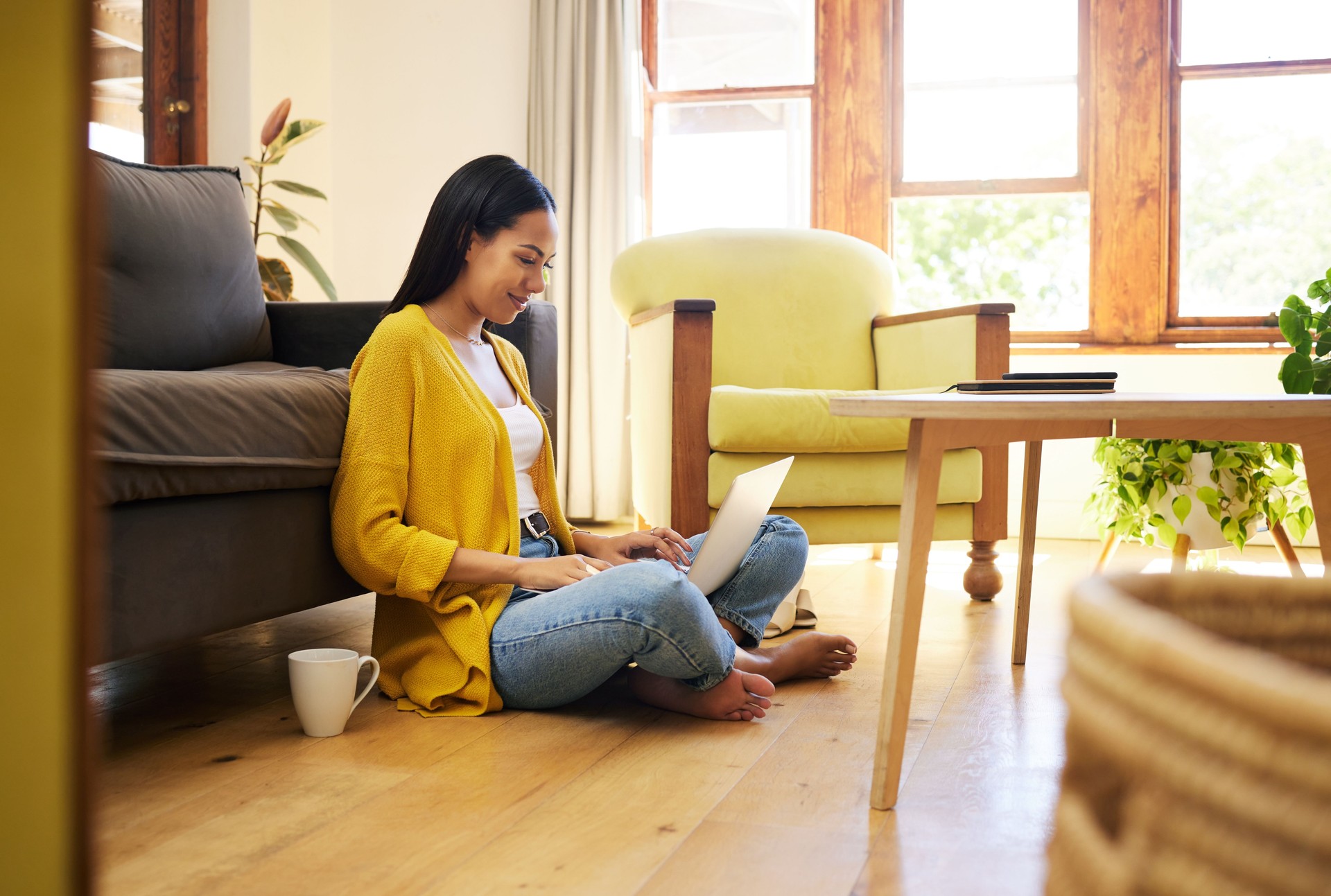Female tenant sitting on the floor with her laptop on her lap and her back against the sofa in a living room. A smiling young female typing on her modern notebook computer at home